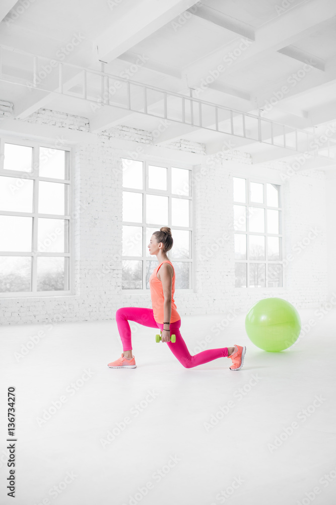 Young woman in colorful sportswear squatting with dumbbels in the white gym with fitness ball on the