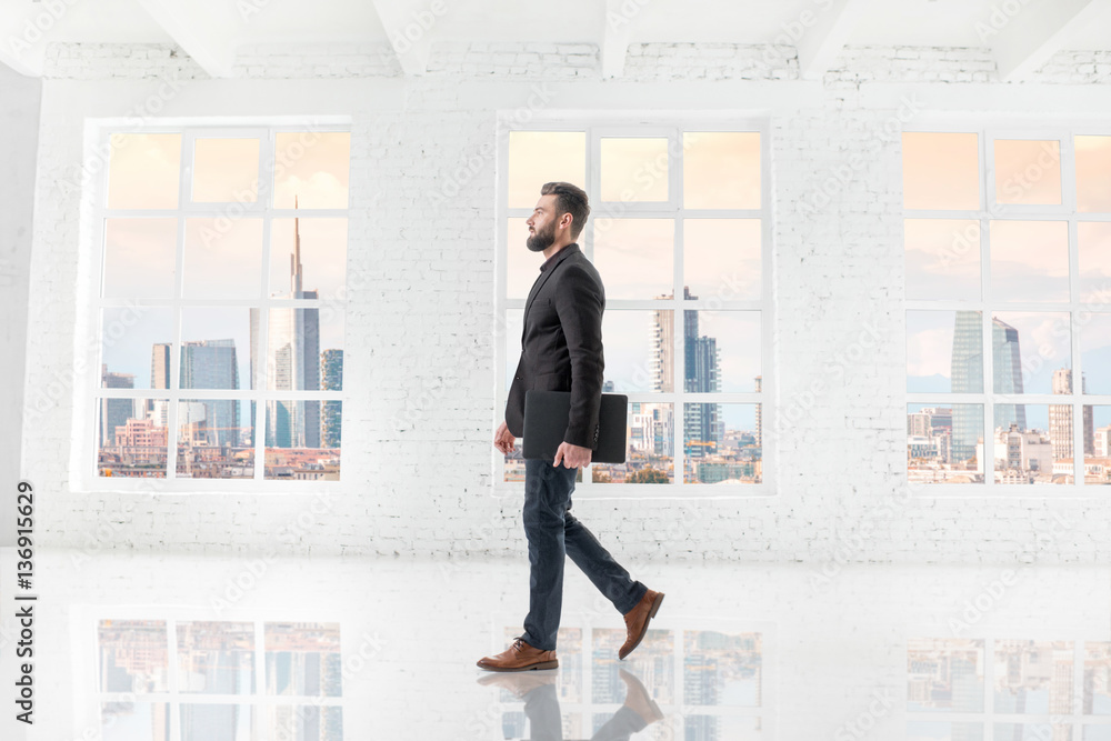 Businessman walking with laptop at the white office interior with big windows with view on the busin