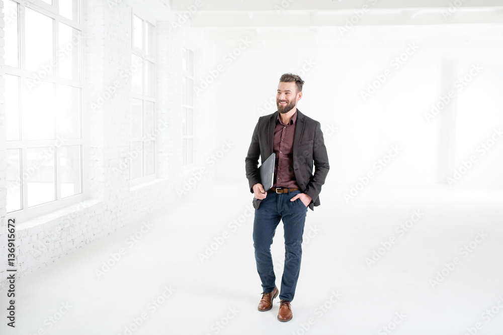 Elegant businessman in the dark suit walking with laptop in the white office interior