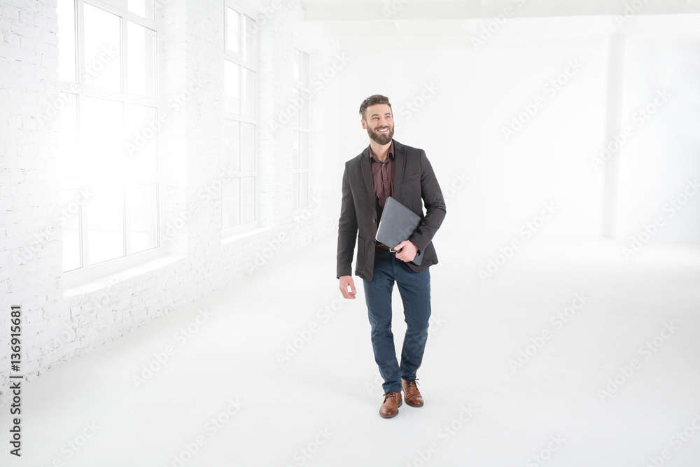 Elegant businessman in the dark suit walking with laptop in the white office interior