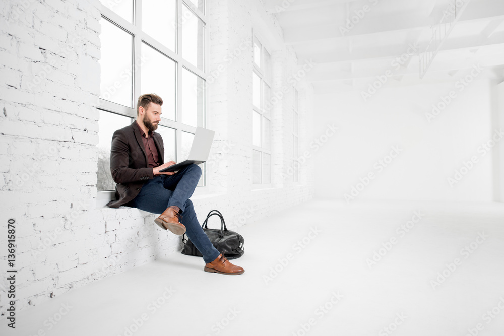 Elegant businessman sitting on the window with laptop in the white office interior