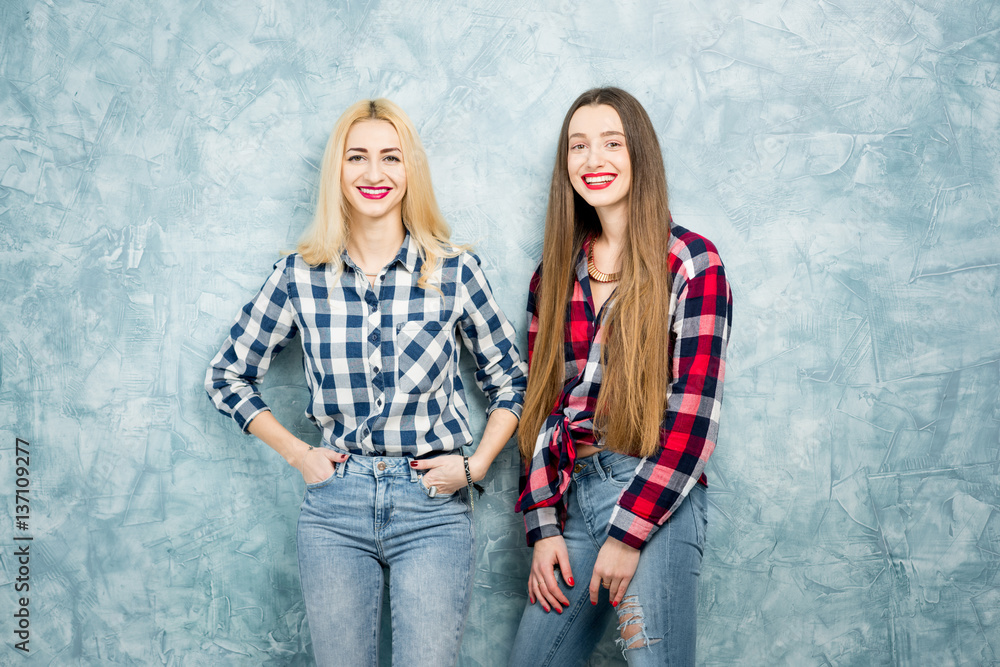 Portrait of two female friends in checkered shirts and jeans together on the blue painted wall backg