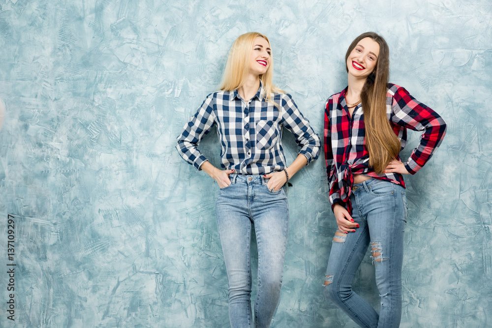 Portrait of two female friends in checkered shirts and jeans together on the blue painted wall backg