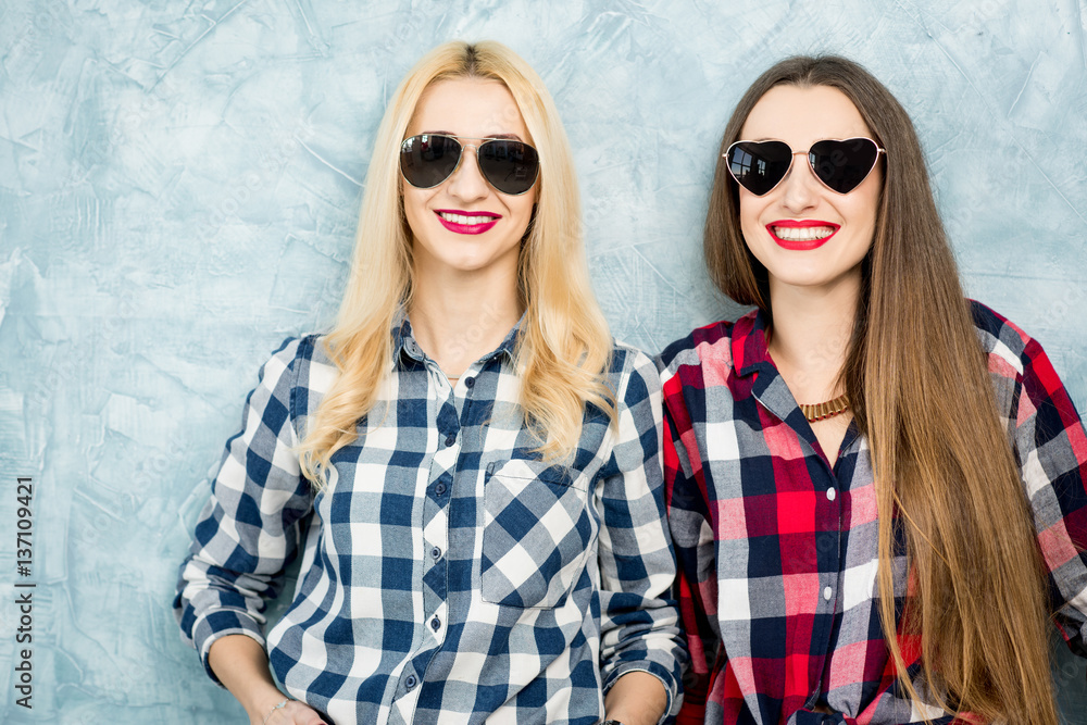 Close-up portrait of two female friends in checkered shirts, jeans and sunglasses on the blue painte