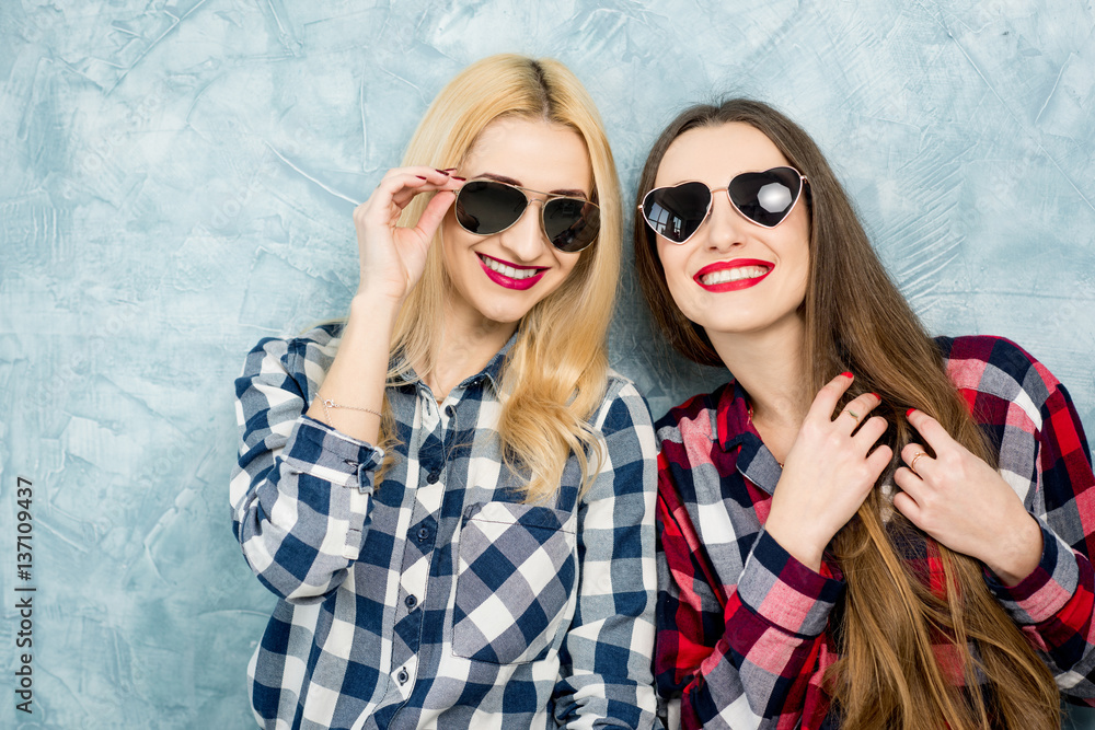 Close-up portrait of two female friends in checkered shirts, jeans and sunglasses on the blue painte