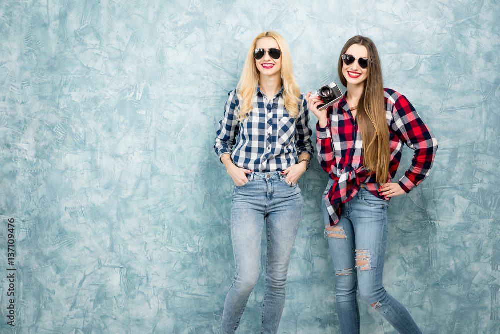 Portrait of two female friends in checkered shirts, jeans and sunglasses on the blue painted wall ba