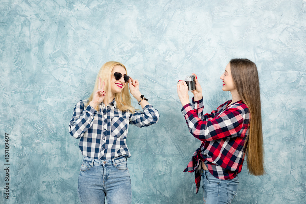 Two female friends in checkered shirts and jeans photographing with retro camera on the blue painted