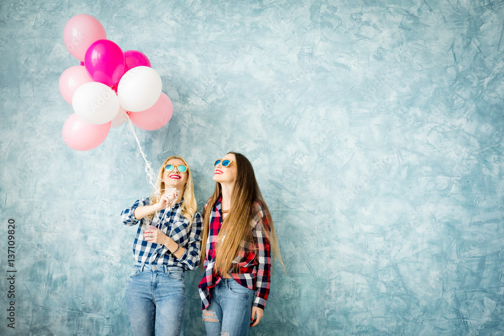 Two female friends in checkered shirts having fun with air balloons on the blue wall background