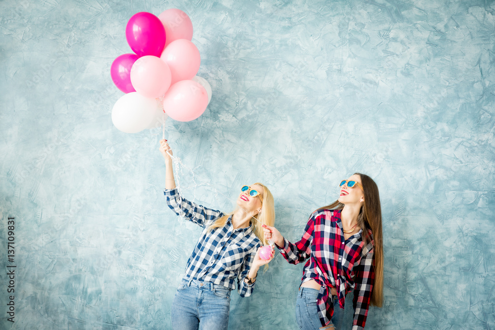 Two female friends in checkered shirts having fun with air balloons on the blue wall background