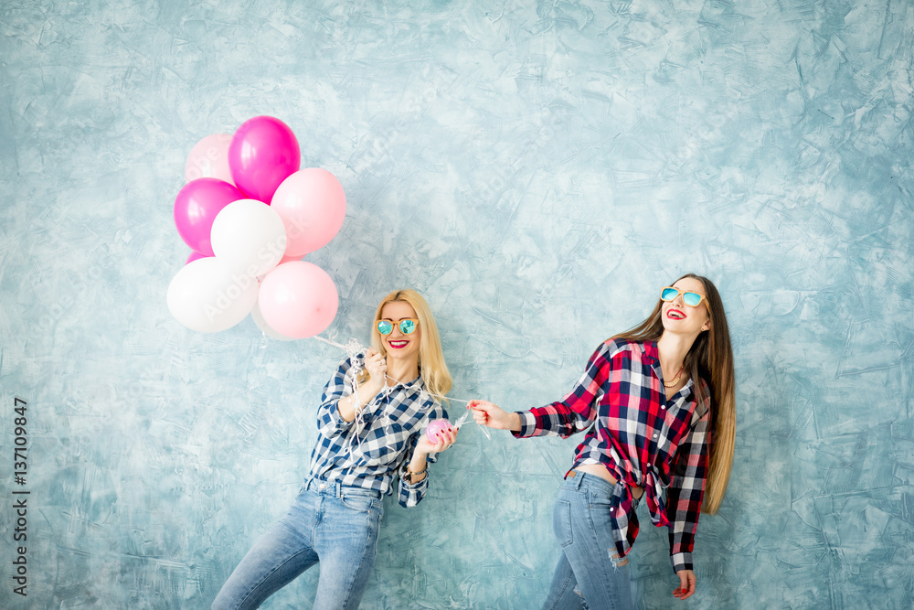 Two female friends in checkered shirts having fun with air balloons on the blue wall background