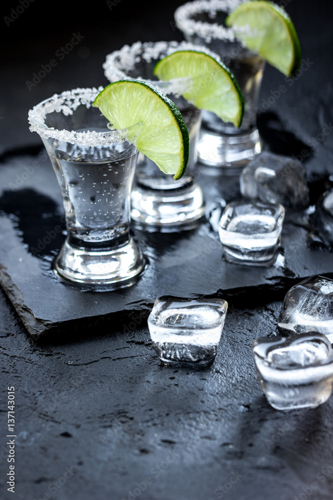 Silver tequila shots with ice and lime on black table background