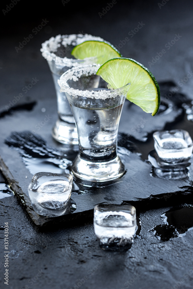 Silver tequila shots with ice and lime on black table background