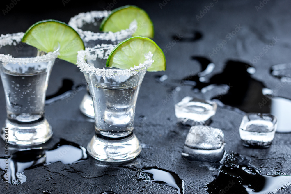 Silver tequila shots with ice and lime on black table background