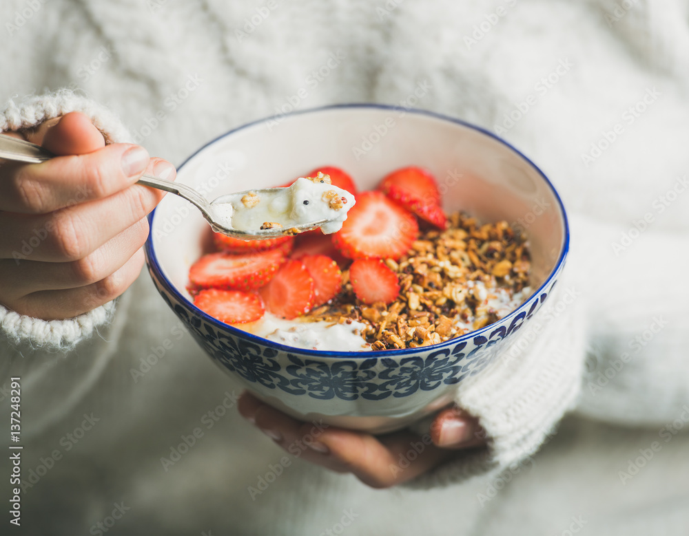 Healthy breakfast greek yogurt, granola and strawberry bowl in hands of woman wearing white loose kn