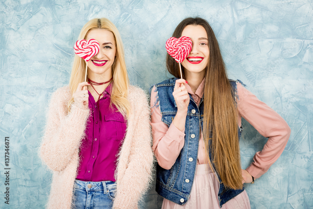 Female stylish friends having fun with candy on the blue wall background