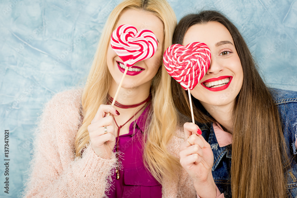 Female stylish friends having fun with candy on the blue wall background