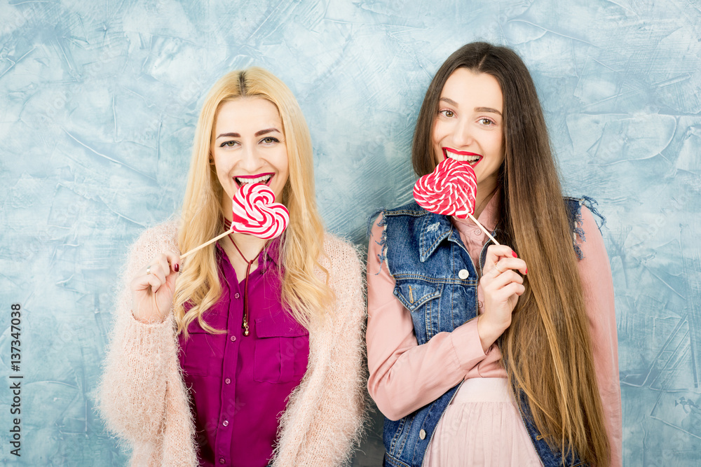 Female stylish friends having fun with candy on the blue wall background