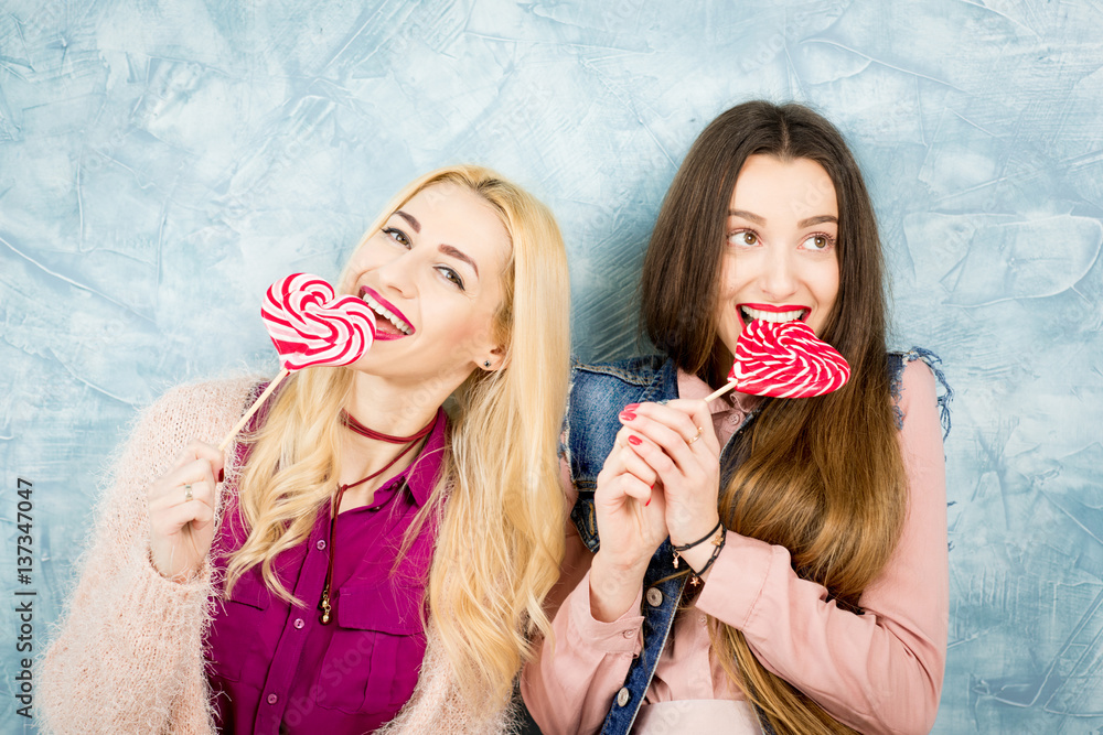 Female stylish friends having fun with candy on the blue wall background