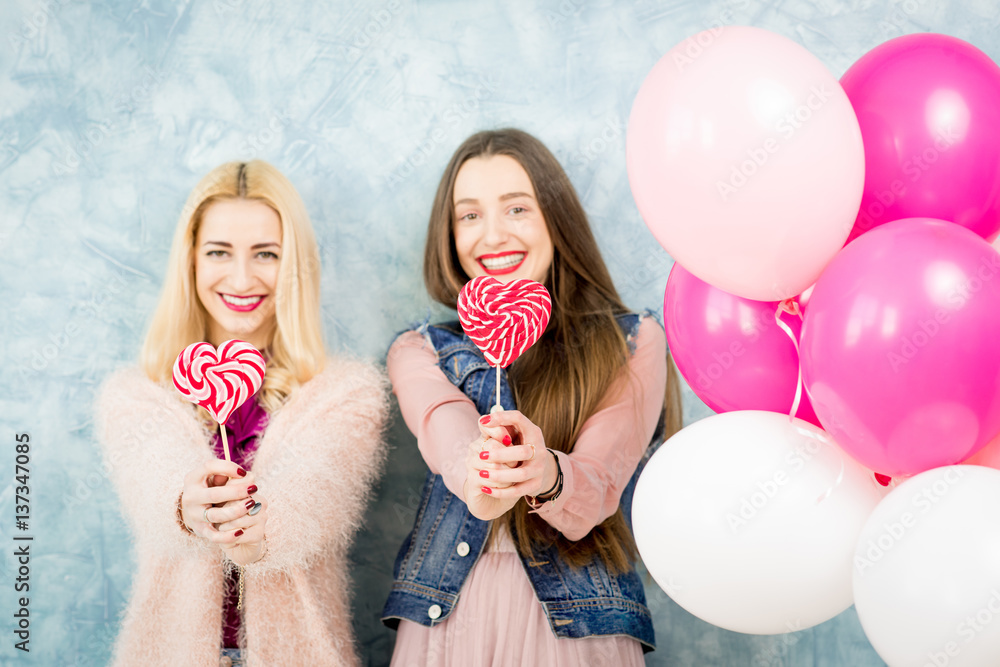 Female stylish friends having fun with candy and baloons on the blue wall background