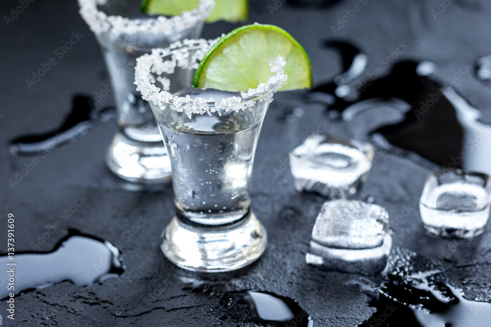 Silver tequila shots with ice and lime on black table background