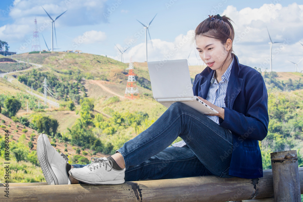 asian women sitting relax on the bamboo and play white laptop computer in the hills and wind turbine