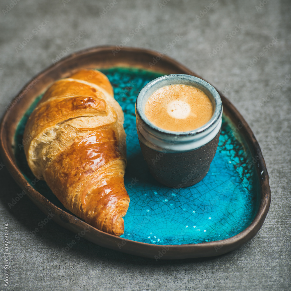 Cup of espresso coffee and croissant in turquoise blue ceramic tray over grey concrete background, s