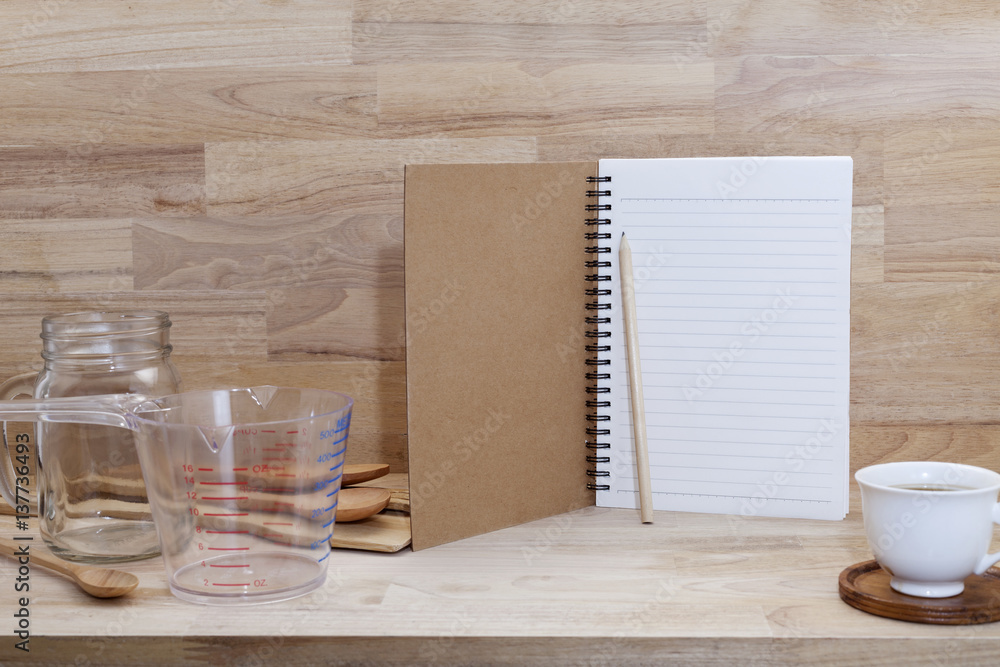 blank notebook with coffice cup and measuring cup and wooden spoon, cooking supplies on wood backgro