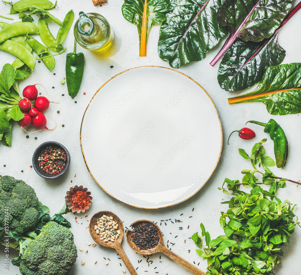 Fresh raw greens, vegetables and grains over light grey marble kitchen countertop, wtite plate in ce