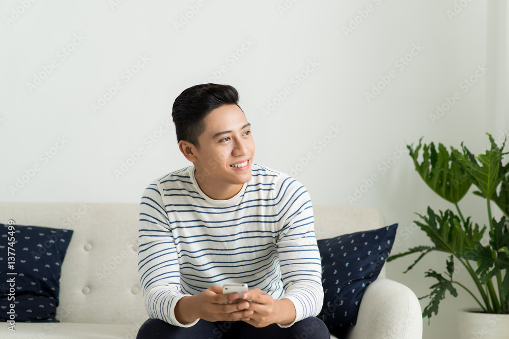 Young handsome asian man using modern mobile smart phone sitting on a couch at home