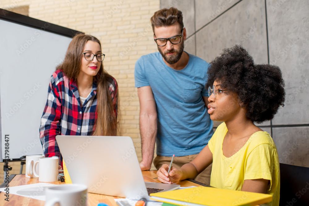 Multi ethnic coworkers dressed casually in colorful clothes talking together sitting with laptop ind