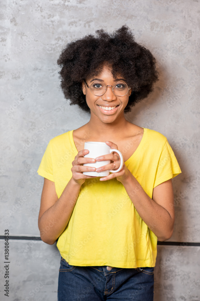 Portrait of a beautiful african woman in yellow t-shirt drinking coffee near the gray wall indoors