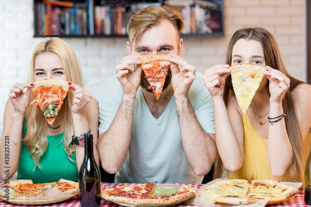 Portrait of young funny friends dressed casually in colorful t-shirts holding slice of pizza at home