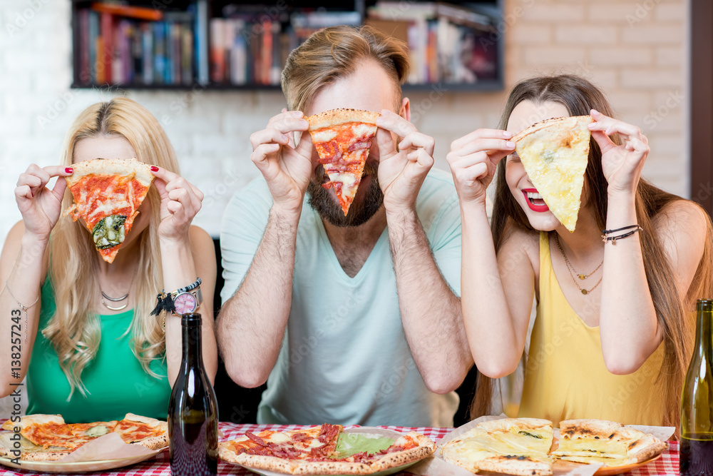 Portrait of young funny friends dressed casually in colorful t-shirts holding slice of pizza at home