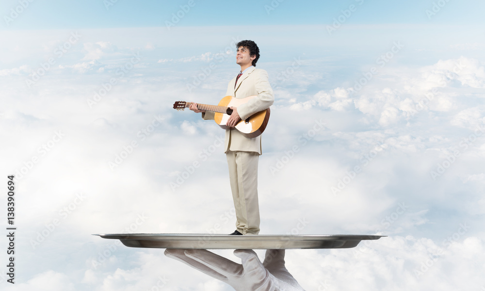 Businessman on metal tray playing acoustic guitar against blue sky background