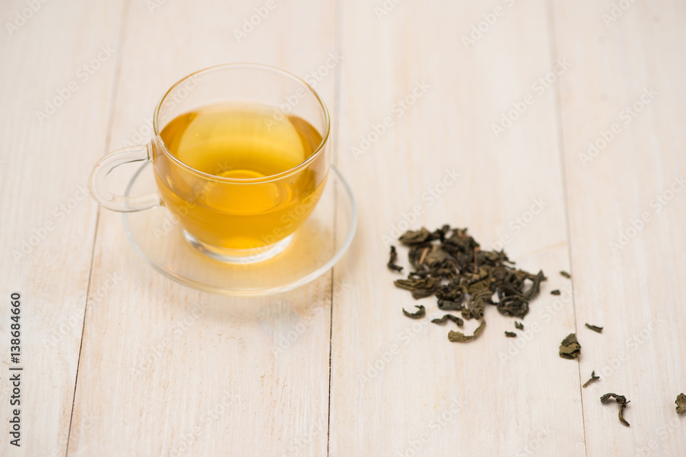 Black tea in a glass cup and tea leaves in wooden spoon on white wooden background.