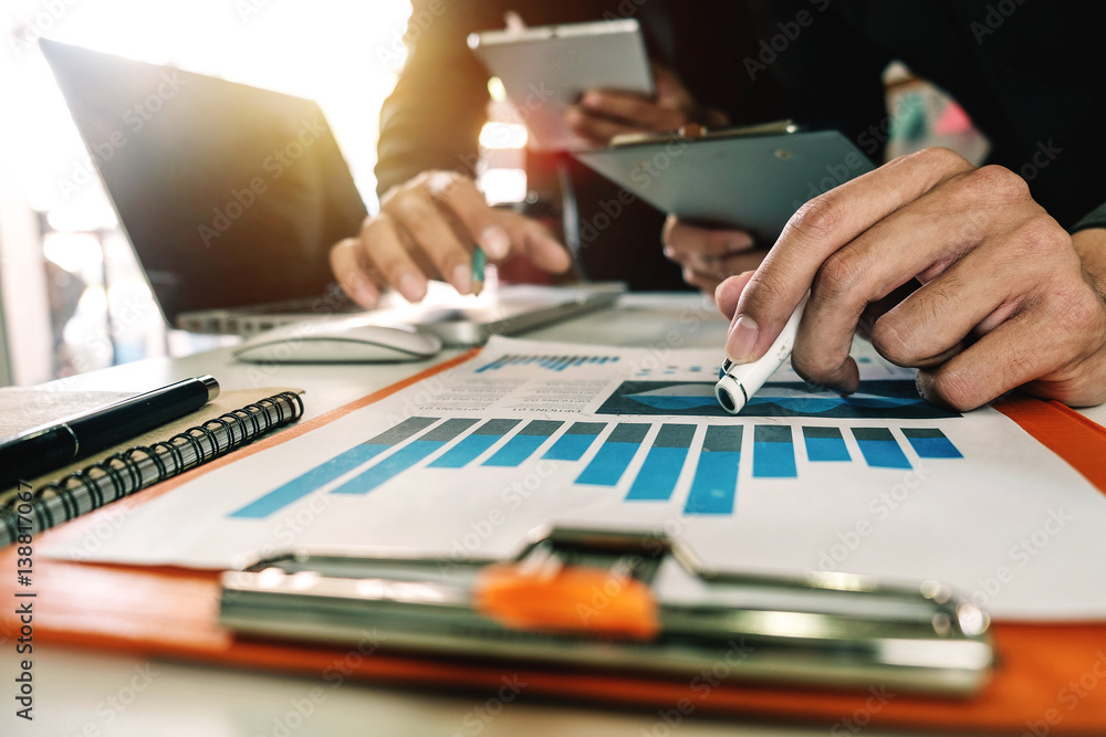 business documents on office table with tablet, smart phone and laptop and two colleagues discussing