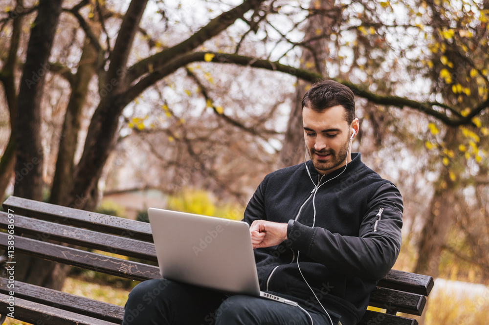 Handsome young man looking at watch while working in the city park