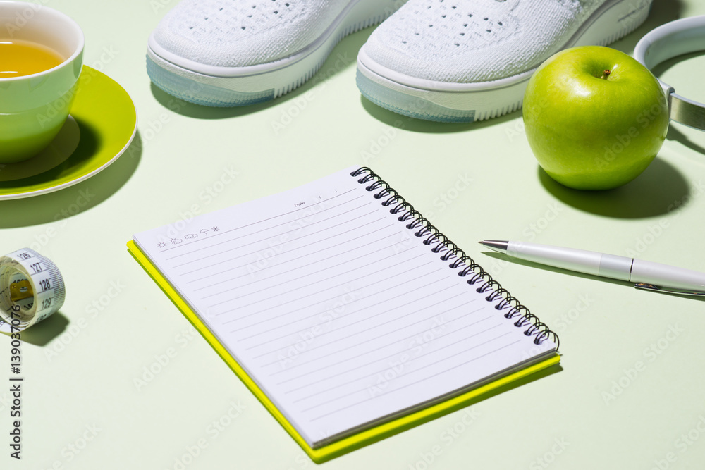 Fitness plan concept. Sneakers, tea, apple and headphone on pastel color background with open notebo