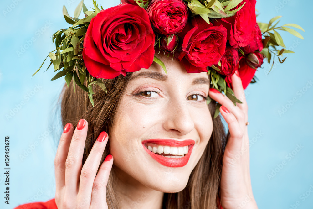 Close-up portrait of a beautiful woman in red dress with wreath made of red roses on the blue backgr