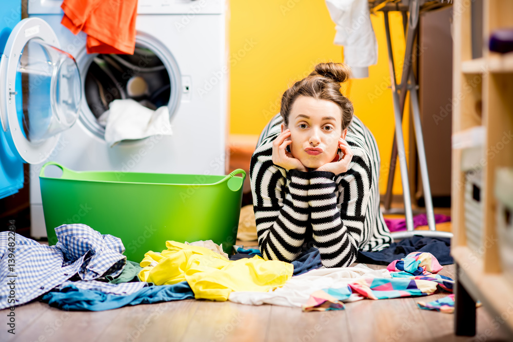 Tired housewife lying on the floor with lots of clothes near the washing machine at home