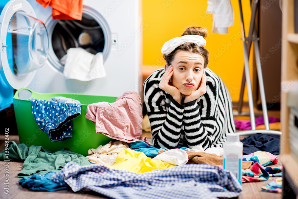 Unhappy housewife sitting with socks near the washing machine with colorful clothes on the floor at 