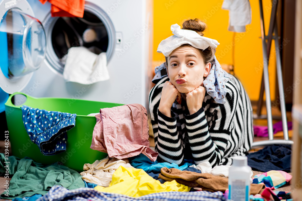 Unhappy housewife sitting with socks near the washing machine with colorful clothes on the floor at 