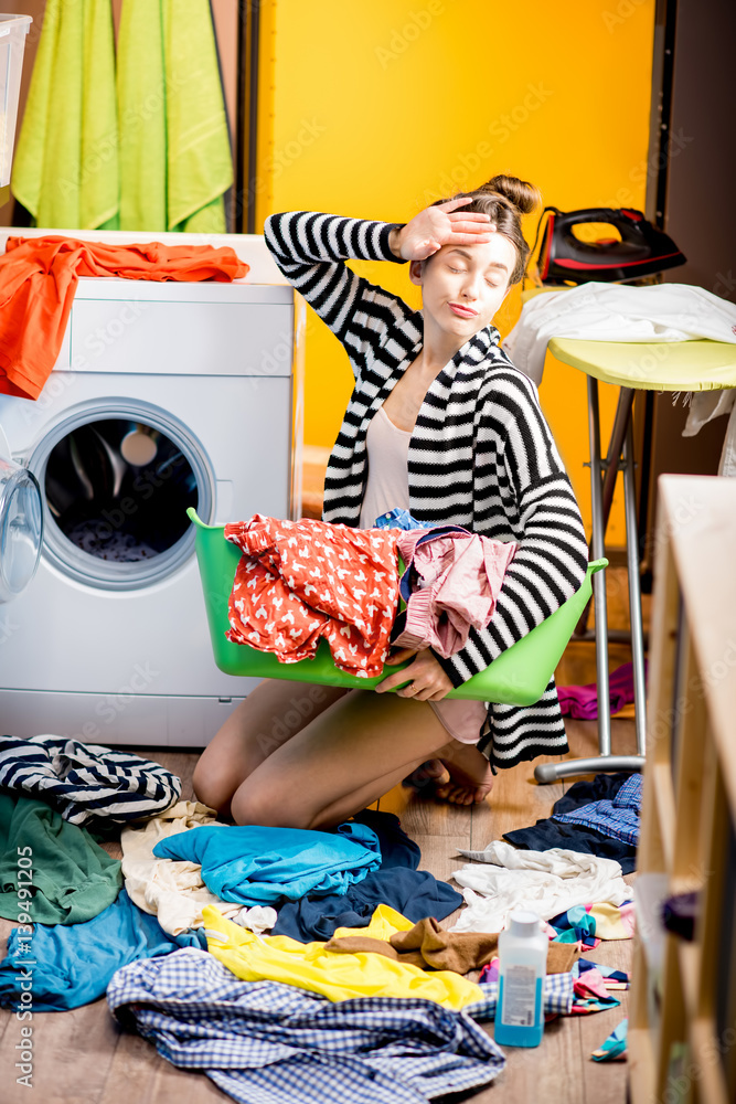 Young housewife holding a basket with clothes near the washing machine sitting on the floor at home