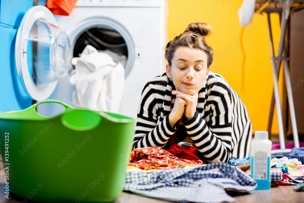 Tired housewife sitting on the floor near the washing machine with colorful clothes at home