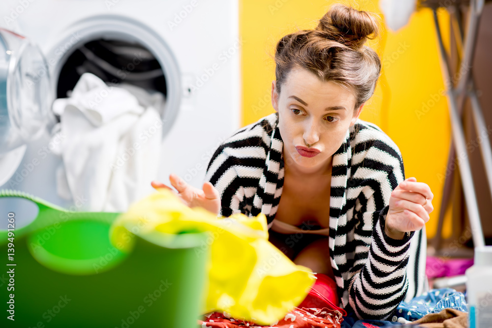 Tired housewife sitting on the floor near the washing machine with colorful clothes at home