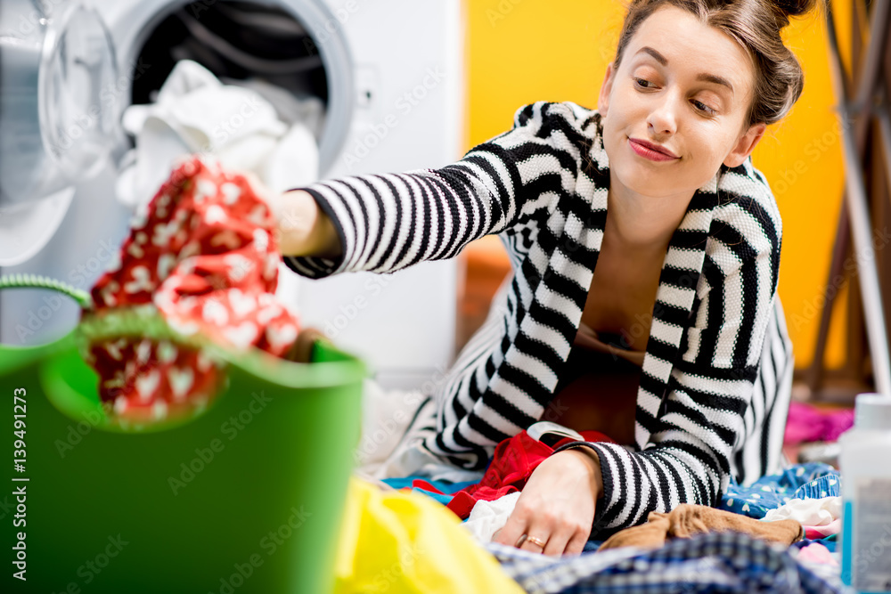 Tired housewife sitting on the floor near the washing machine with colorful clothes at home