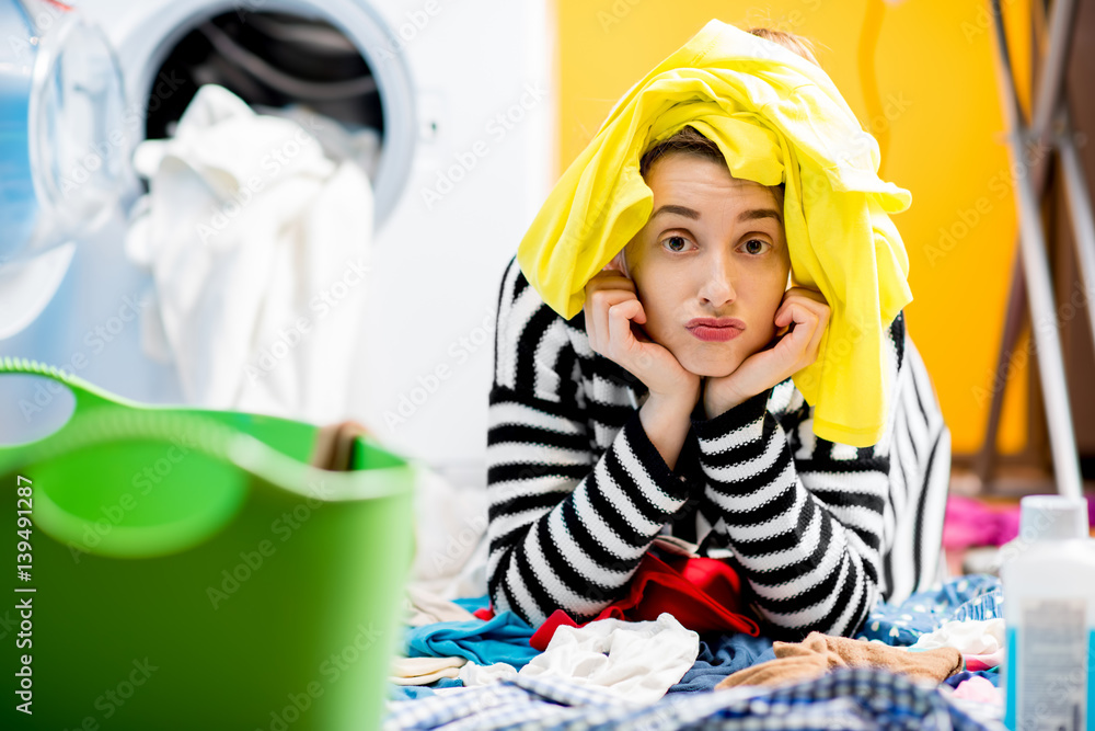 Tired housewife sitting on the floor near the washing machine with colorful clothes at home