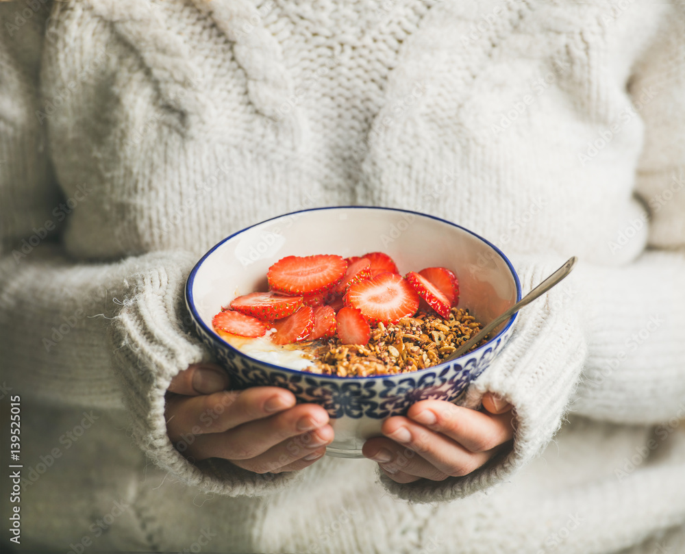 Healthy breakfast greek yogurt, granola and strawberry bowl in hands of woman wearing white loose wo