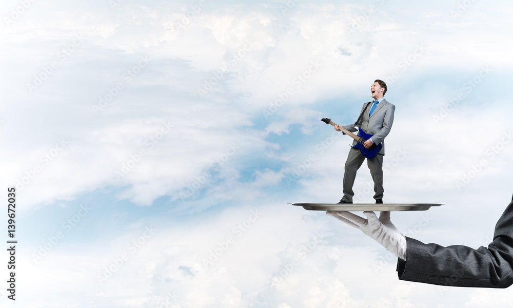 Businessman on metal tray playing electric guitar against blue sky background