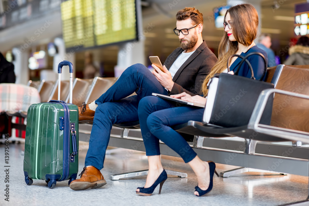 Elegant business couple sitting with phone and book at the waiting hall in the airport. Business tra